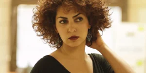 Close-up of a woman with curly hair and teal earrings, hand on neck, looking to the side in soft indoor light.