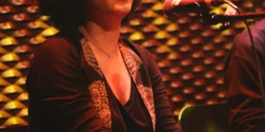 Close-up of the seated vocalist singing into a mic at the WUK Women’s Day concert, lit by warm red–gold stage lights against a textured patterned wall, hands folded on her lap.