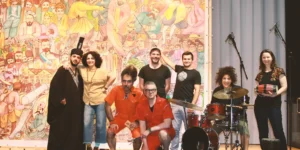 Behind-the-scenes group photo for Shahid: performers and musicians pose on stage in front of a colorful Persian-style mural—one in a black robe with a tall hat, two in orange jumpsuits crouching, a drummer at a kit, and another holding a hand drum—with stage microphones visible.