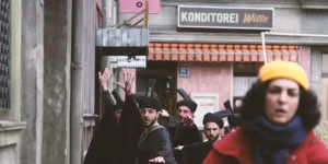 Film still from Shahid: in a narrow alley, a woman in a yellow beanie and red coat moves past with a bicycle in the foreground while a robed chorus with tall black headpieces advances behind her with raised hands; shop signs (“KONDITOREI,” “CASINO”) line the street.