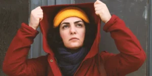 Film still from Shahid: a woman in a yellow beanie and red coat raises her hood while standing before an ornate gray door, looking upward with a determined expression.