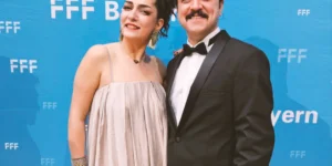 Red carpet photo related to Shahid: actors Baharak Abdolifard and Nima Nazarinia pose before a blue “FFF Bayern” step-and-repeat—one in a light, flowing gown with a small black clutch, the other in a black tuxedo with bow tie, both smiling at the camera.
