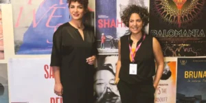 Festival photo: two guests stand in front of a wall of film posters—including the Shahid poster—both dressed in black; one wears a lanyard and smiles toward the camera.
