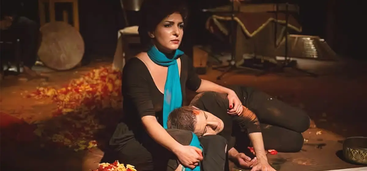 Baharak Abdolafard cradles a fellow performer’s head on her lap during “Siavoshan” (October 2015), seated amid scattered rose petals on a dim stage with a frame drum and santur in the background.
