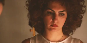 Film still: close-up of a curly-haired actress, Baharak Abdolifard, in a white blouse, pensive under soft studio lighting.