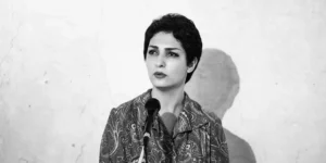 Black-and-white portrait of the vocalist facing forward behind a microphone, light wall backdrop with her shadow visible.