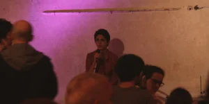Close-up of a vocalist at a microphone against a purple-lit plaster wall, singing as audience heads blur in the foreground; part of a drum kit and musicians at right—live performance for Arash // Heimkehrer.