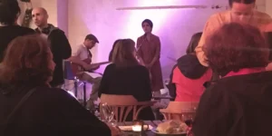 Audience view of live music at Arash // Heimkehrer: a vocalist sings at a microphone under a purple wall wash with a guitarist to the left, while a café-style crowd sits at small tables with candles in the foreground.