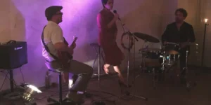 Live music at Arash // Heimkehrer: a vocalist sits with a microphone and tambourine between a seated electric guitarist (left) and a drummer (right) against a purple-lit plaster wall; cables and floor lights, with a table and plate in the foreground.