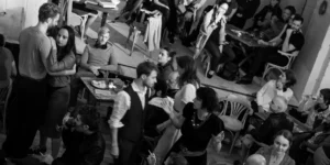 Black and white photo of a theater performance in a café-like setting, where the audience is seated at small tables with drinks and food. In the foreground, a man and woman stand closely together as if dancing, while other performers interact among the audience. The atmosphere is intimate and immersive, blurring the line between stage and spectators.