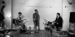 Black-and-white scene of a small indoor concert: guitarist on the left, vocalist centered at a mic stand, drummer on the right; wooden floor and empty chairs in the foreground.