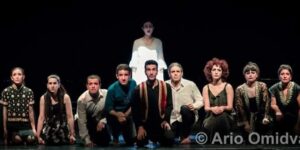 Stage photo from Nodbeh: nine performers kneel in a row under a tight spotlight while a tenth figure stands behind them in white, singing against a black background.