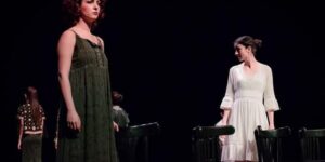 Stage photo from Nodbeh: two performers stand apart among scattered chairs—one in a long green dress facing forward, another in a white dress turned sideways—while others fade into the dark background.