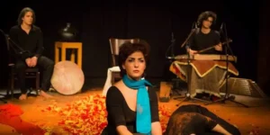 Lead performer sits center stage in a turquoise scarf amid scattered petals, with frame-drum and santur players in the background — scene from the 2015 stage piece “Siavoshan.”