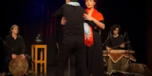 Close embrace during the “Siavoshan” duet, conveying grief and resolve, musicians visible in soft focus upstage.