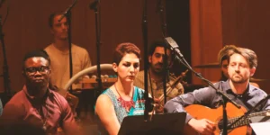Seated vocalist, Baharak Abdolifard between a guitarist and another performer, focused and still; percussion and a string player visible in the background with close microphones.
