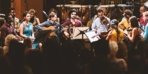 Wide shot of the ensemble on stage before a full audience—vocalist, two guitarists, strings, woodwinds, and percussion arranged in a semi-circle at RadioKulturhaus.