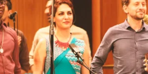 Vocalist, Baharak Abdolifard in a turquoise patterned dress during the curtain call, standing between musicians; microphones and warm wooden panels; audience hands in foreground.