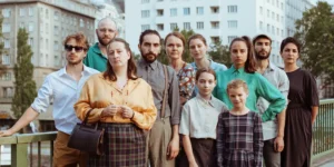 Group portrait for the theatre project Arash: about a dozen performers of different ages stand closely together on a city bridge, facing the camera with neutral expressions; apartment blocks and trees in the background.