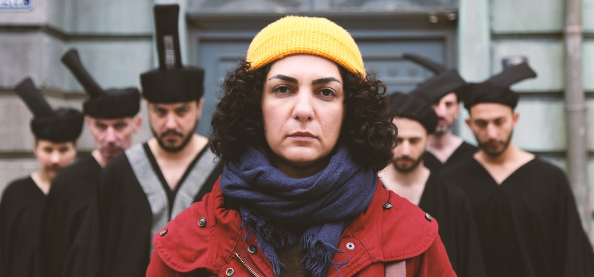 from the film Shahid: a woman, Baharak Abdolifard, in a yellow beanie, red coat, and blue scarf looks straight at the camera, while a group of robed performers in black with tall headpieces stand blurred behind her on a city street.