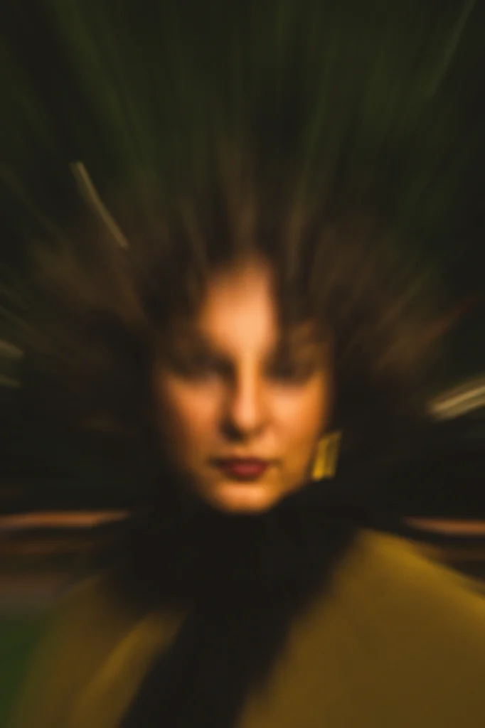 Abstract zoom-blur portrait of a woman with curly hair and red lipstick, wearing a dark scarf and olive coat, centered against a dark green outdoor background.