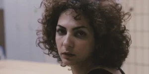 Trailer still from The Photographer, His Lover, His Sister and Her Lover (2017): close-up of a curly-haired woman turning toward the camera, soft light and tiled wall in the background, pensive expression.