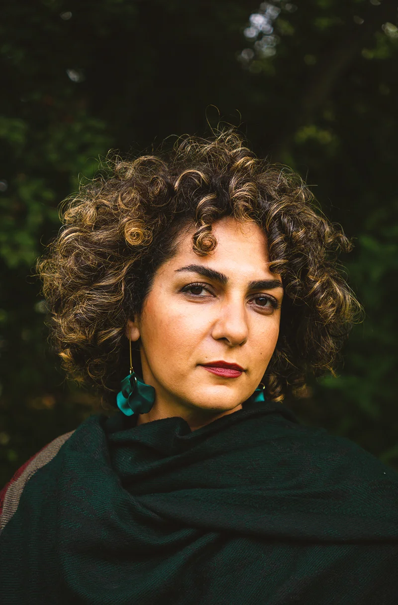 Portrait of Baharak Abdolifard with short curly hair, teal earrings, and a dark green shawl, standing outdoors against leafy background.
