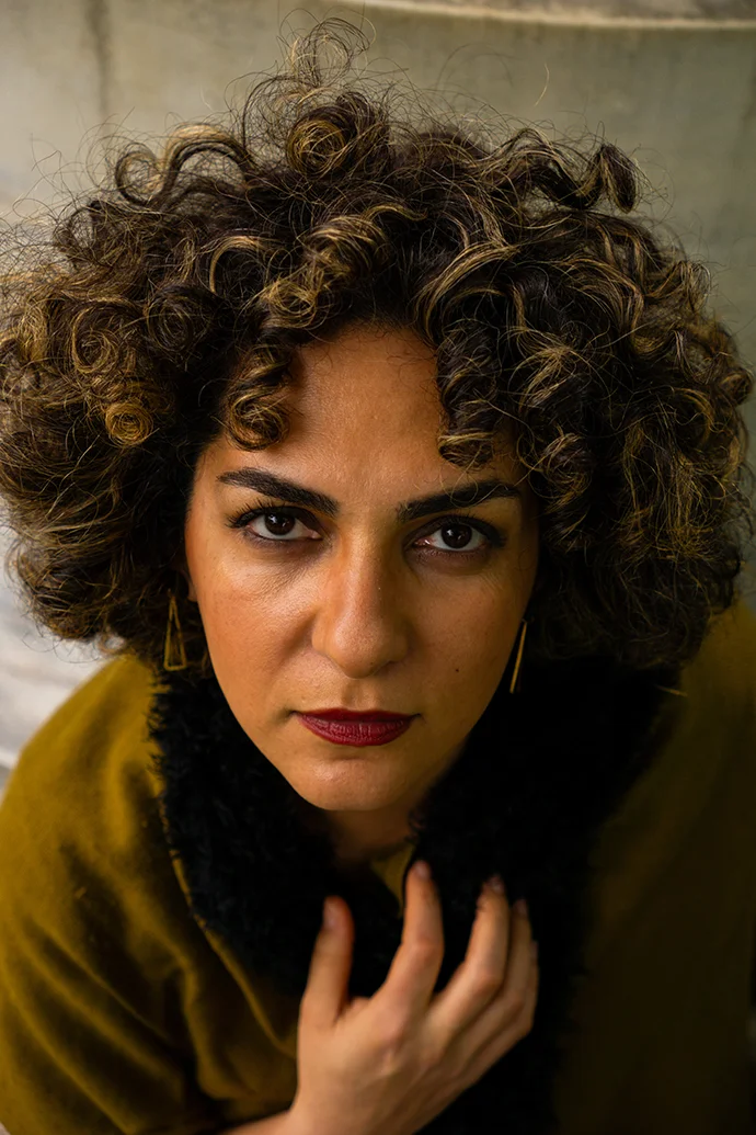 Close-up portrait of a curly-haired woman with red lipstick, hand at collar, looking directly at the camera.